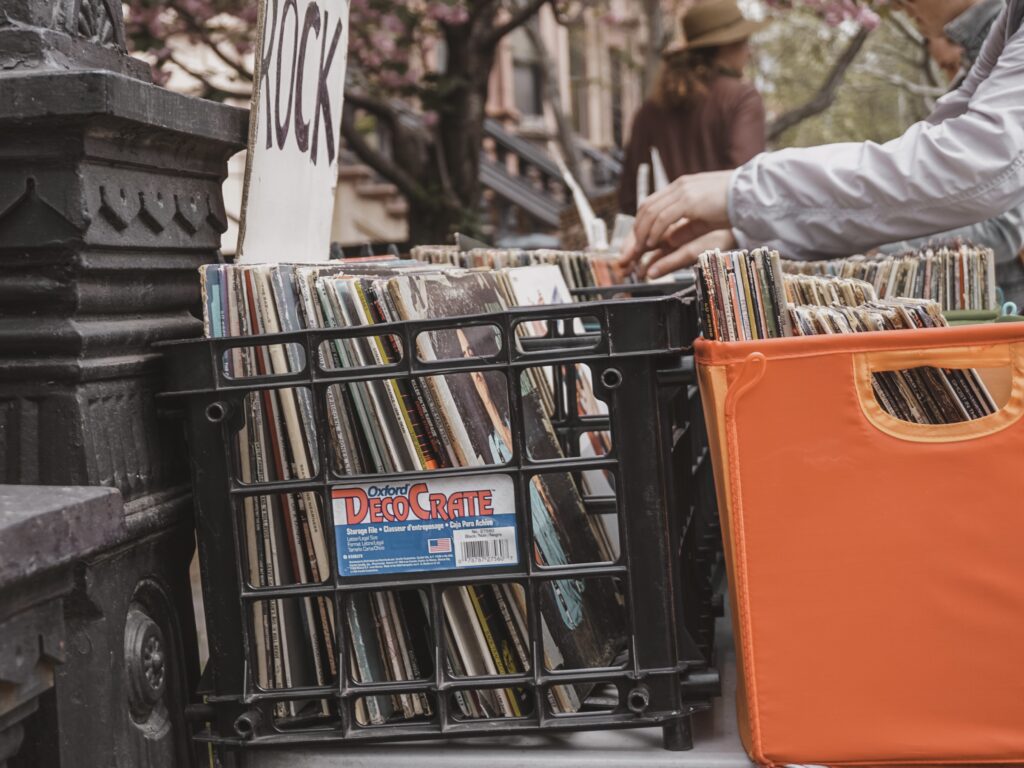 Foto com caixas de discos de vinil à venda em rua de Nova Iorque, close em caixa com a sinalização “rock” e em uma mão que mexe nos discos.