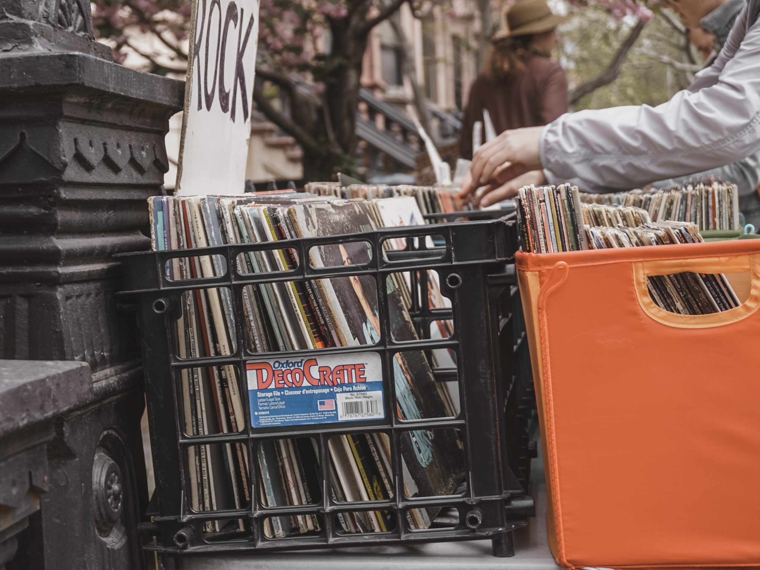 Foto com caixas de discos de vinil à venda em rua de Nova Iorque, close em caixa com a sinalização “rock” e em uma mão que mexe nos discos.